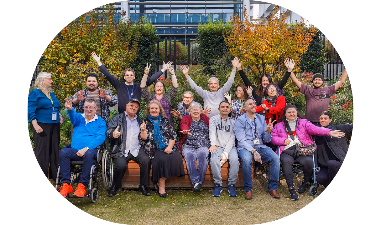 Group photo of BrainLink clients, carers, and staff smiling and raising their arms outdoors, celebrating community and support for people living with brain injury.